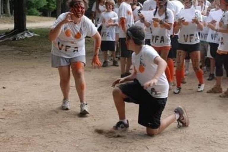 A group of people in white shirts with painted faces play a game outdoors; one person kneels in front.