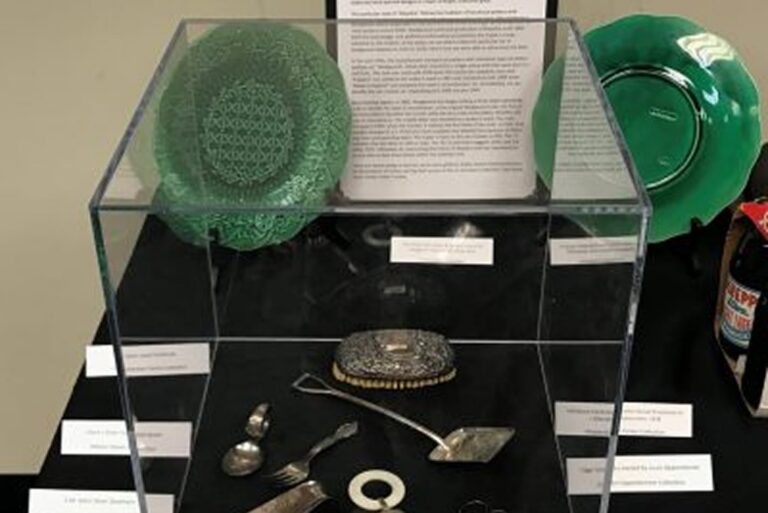 A glass case displays vintage silverware, a brooch, green plates, and historical information cards behind and beside it.