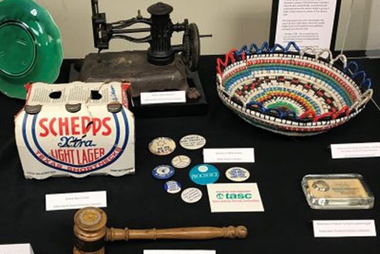 A display with vintage items: beer packaging, sewing machine, basket, buttons, gavel, and business cards on a black table.