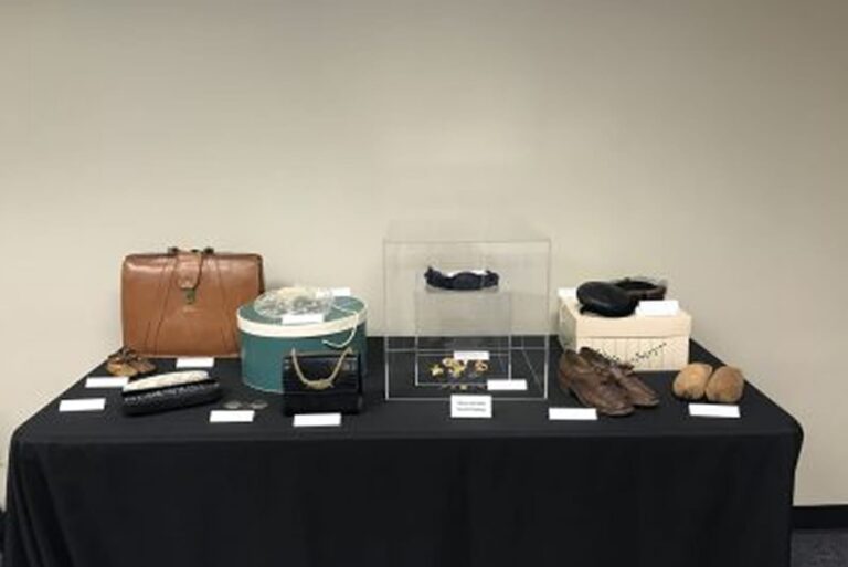 A display table with vintage accessories, including bags, shoes, hats, and jewelry, set against a plain wall.