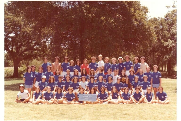 A large group in blue shirts poses outdoors on grass with trees behind them, holding a sign in the front row.