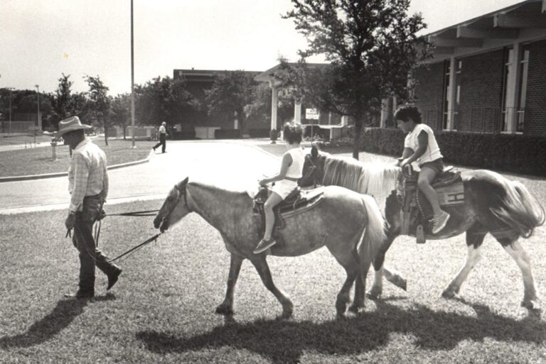 A man leads two children riding ponies on a grassy area near school buildings on a sunny day.