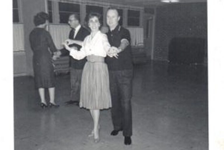 Two couples dance in a spacious room with wooden floors and large windows, dressed in mid-20th century clothing.