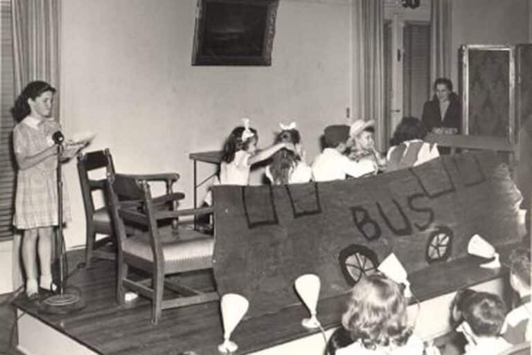 Children perform a school bus-themed play on stage, with a girl speaking at a microphone and an audience watching.
