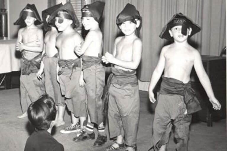 Six children in costumes stand in a line indoors while an adult crouches in front of them, watching.