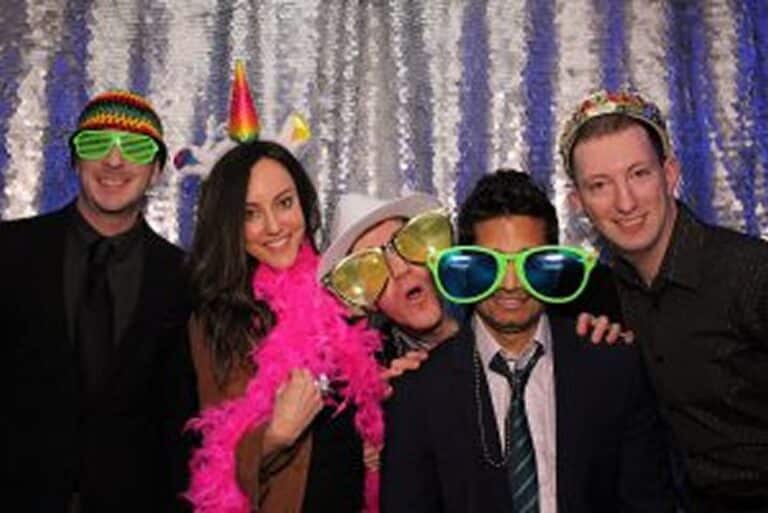 Five people in fun costumes and props, posing and smiling in front of a shiny silver sequin backdrop.