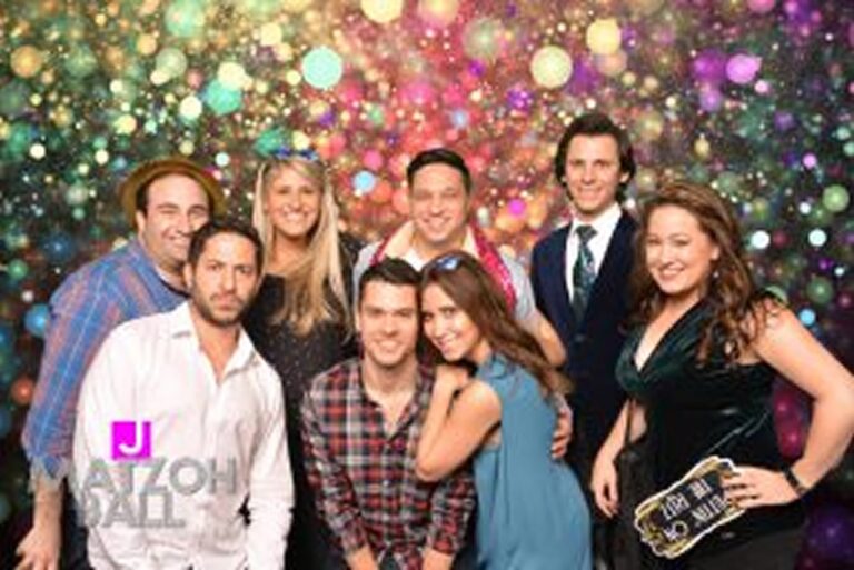 Group of nine adults posing and smiling in front of a colorful, sparkly backdrop at a party or event.