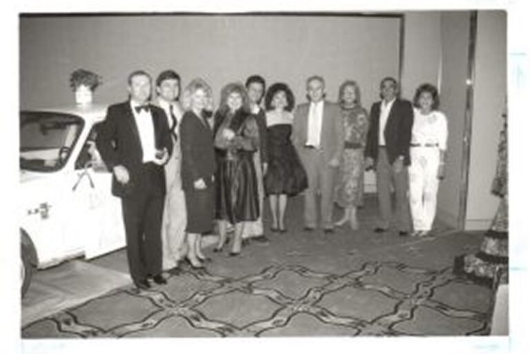 A group of ten people dressed formally stand together indoors next to a car, posing for a photo.