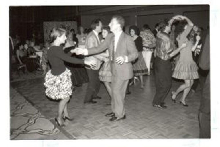 People in vintage attire dance in pairs on a patterned floor at an indoor social event or dance party.