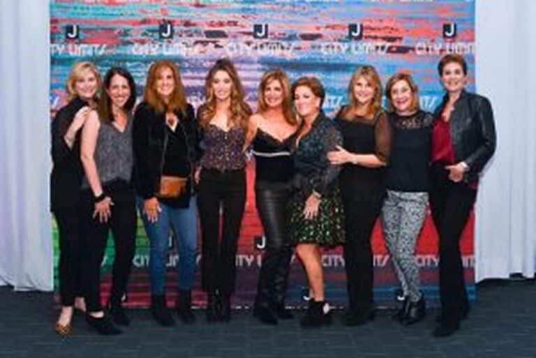 Eight women posing together in front of a colorful, branded backdrop, smiling for the camera.