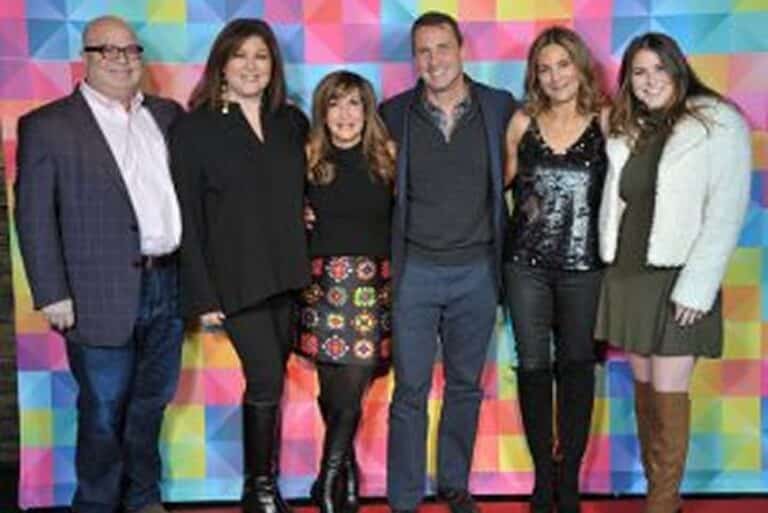 Six people stand in a row, smiling, in front of a colorful geometric-patterned backdrop.