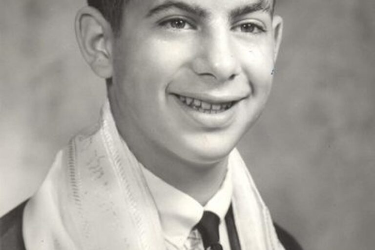 Black and white portrait of a smiling boy wearing a suit, tie, and a prayer shawl over his shoulders.