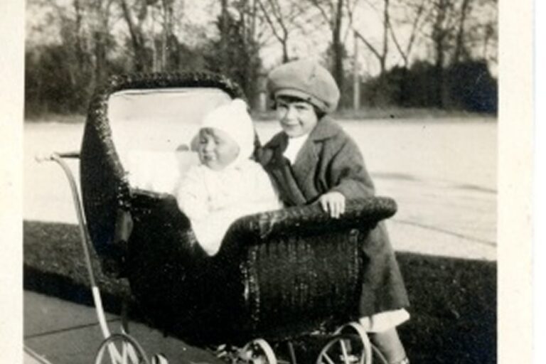 Two children outside: a baby in a vintage stroller and an older child standing beside, both dressed warmly.