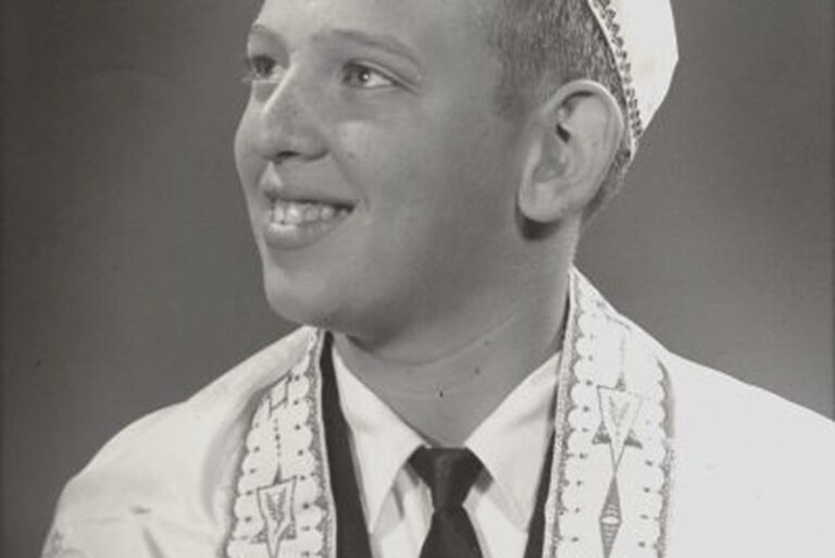 Smiling boy wearing a yarmulke and prayer shawl, dressed in a suit and tie, looking off to the side.