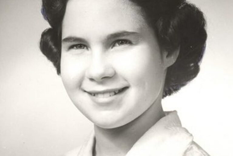 Black and white portrait of a young woman smiling, with dark wavy hair and wearing a light-colored collared shirt.