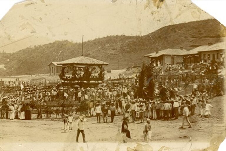 Sepia photo of a large crowd gathered outdoors near buildings and hills, possibly for a festival or public event.