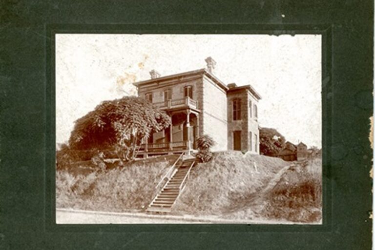 Old two-story brick house on a grassy hill, with stairs leading up to a covered porch and large vines on one side.