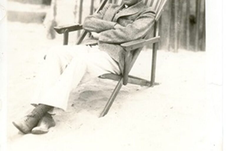 Person sitting in a wooden chair on sand, dressed in a suit jacket and trousers, arms crossed, looking relaxed.