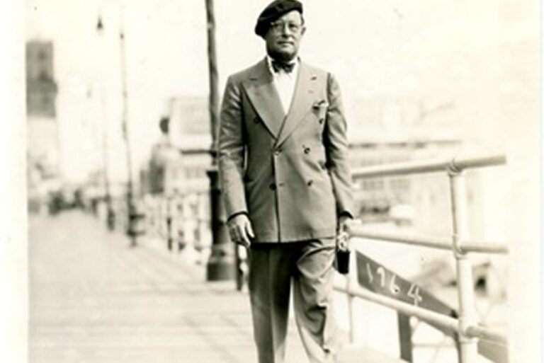 Man in a double-breasted suit and beret walking along a boardwalk or pier, with a blurred city background.