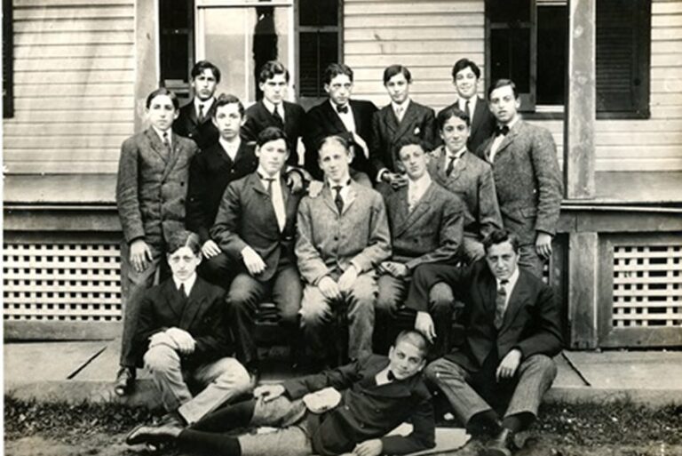 Fifteen young men in suits pose for a formal group photo in front of a wooden building.
