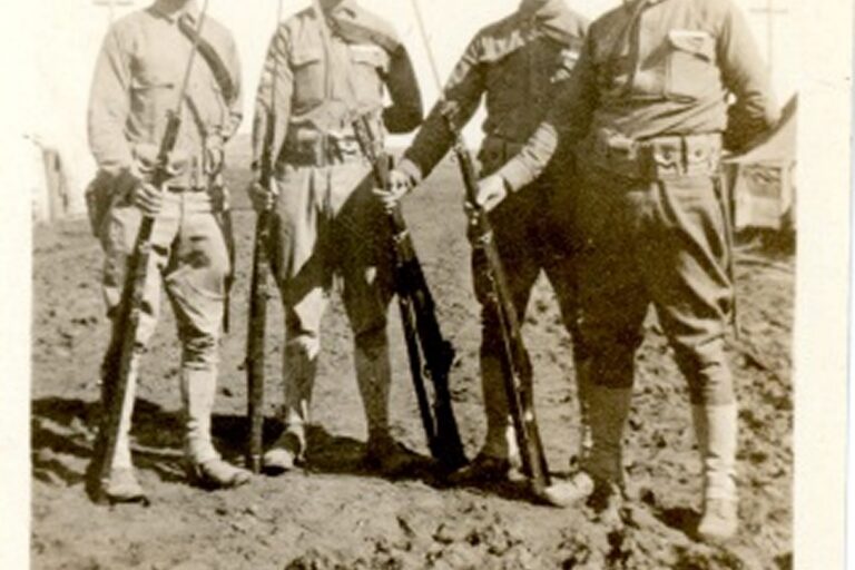 Four soldiers in uniform stand on dirt, holding rifles, posing for a photo in daylight.