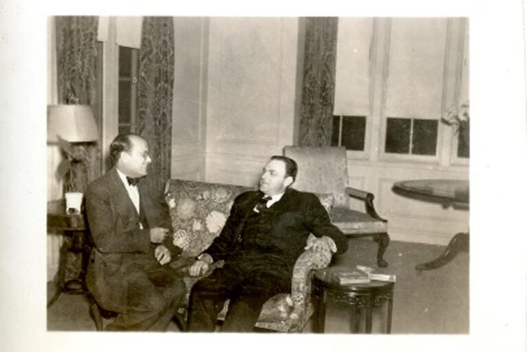 Two men in suits sit and talk in a warmly lit, elegant room with couches, a table, and large curtained windows.