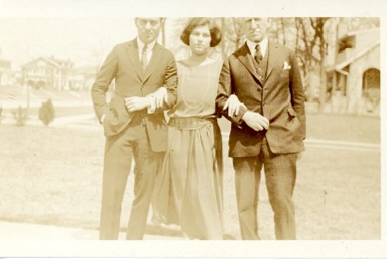 Three people in early 20th-century attire stand arm in arm outside on a sunny day with houses in the background.