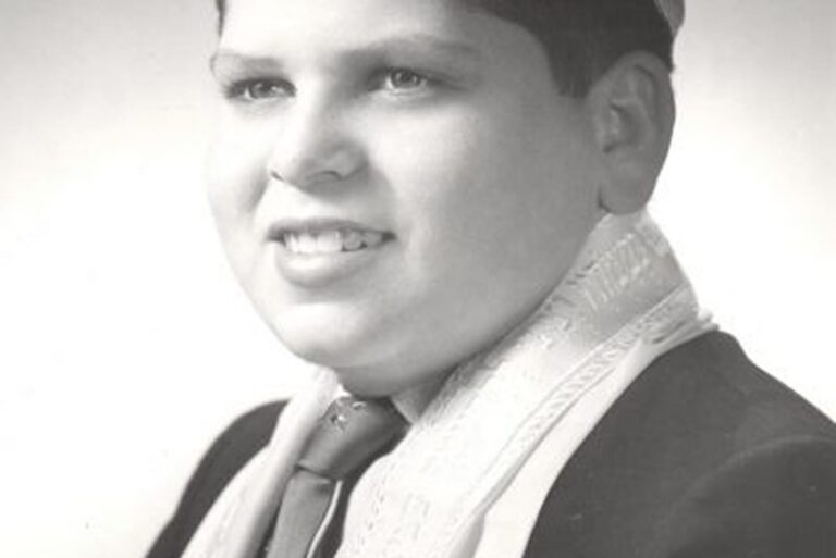 Black and white portrait of a smiling boy wearing a suit, tie, and patterned scarf or sash, facing slightly right.