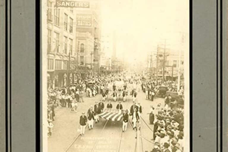 Black-and-white photo of a parade on a city street with people marching and crowds watching on both sides.
