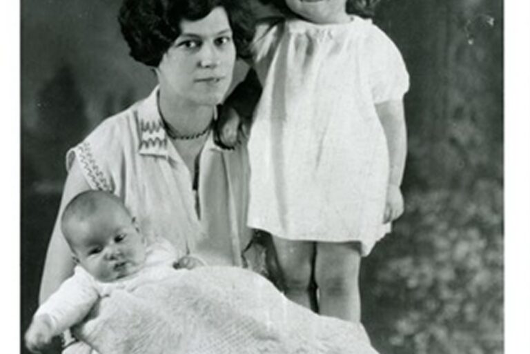 Woman poses with a baby on her lap and a young girl standing beside her, all dressed in light clothing. Black and white photo.