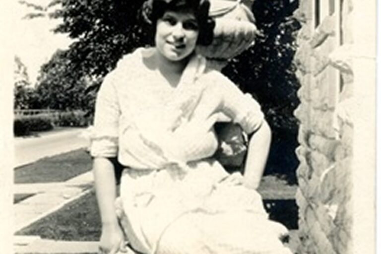 Smiling woman in a light dress poses outdoors by a stone building, with trees and a street visible in the background.