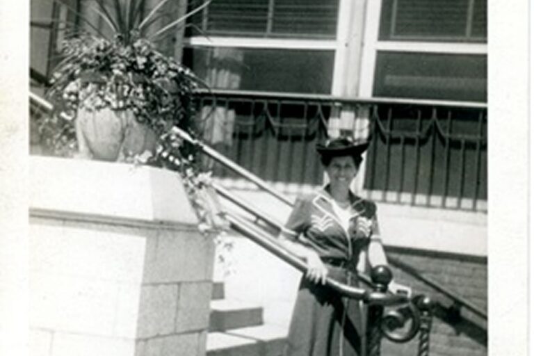 A woman in a dark dress and hat stands by a railing next to a potted plant and stairs outside a building.