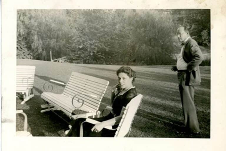 A woman sits on a bench outdoors while a man stands behind her on a grassy lawn, trees in the background.