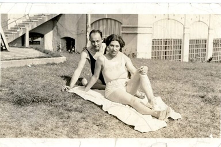 Two people in swimsuits sit on a towel on grass, with buildings and stairs in the background, in a vintage photo.