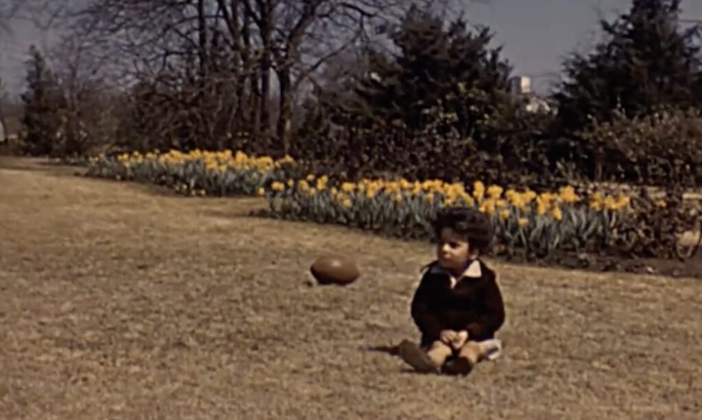 A young child sits on a lawn near a football, with yellow flowers and trees in the background.