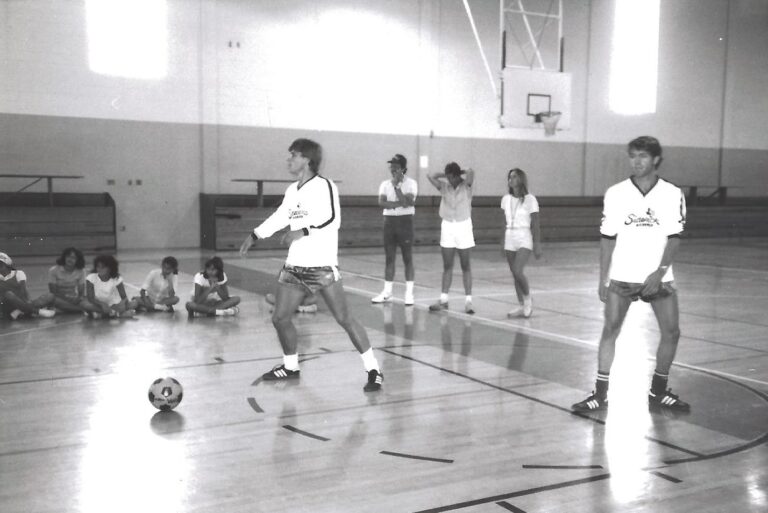 Two people in soccer uniforms in a gym, one about to kick a ball, with others watching and sitting nearby.
