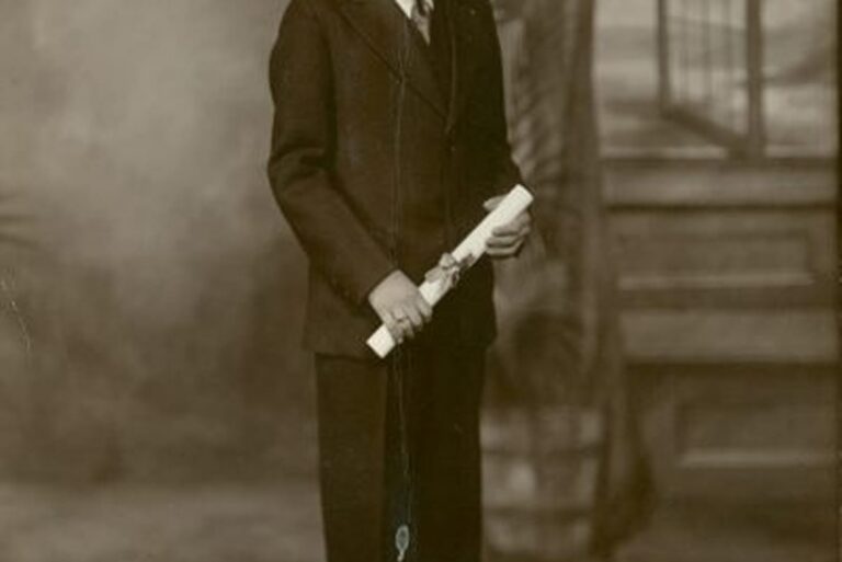 Young man in a suit holding a rolled diploma poses for a formal portrait in a vintage studio setting.
