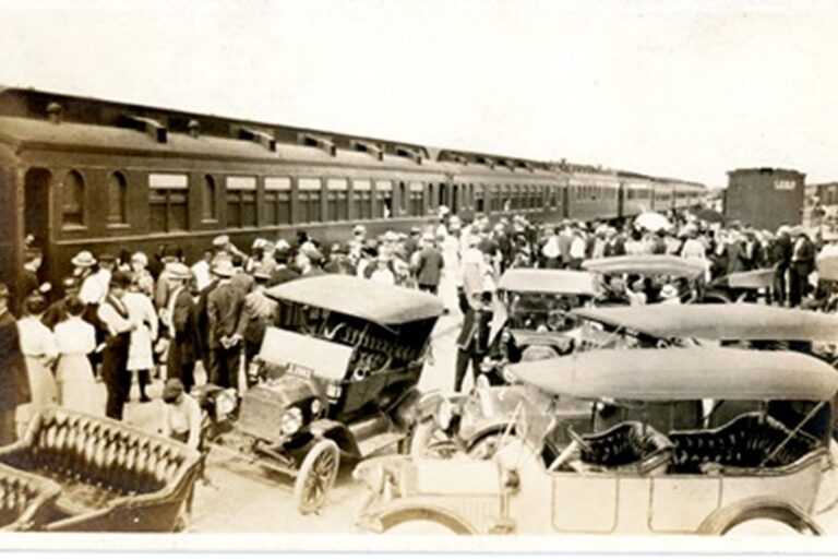 A crowd gathers near vintage cars and a long train at a busy station, dressed in early 20th-century clothing.