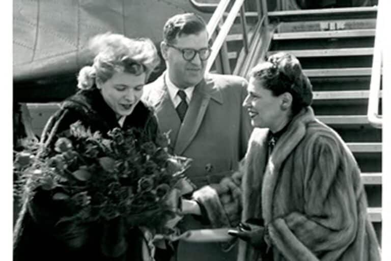 Three adults, two women in fur coats and a man in glasses, stand by airplane stairs. One woman holds a bouquet.