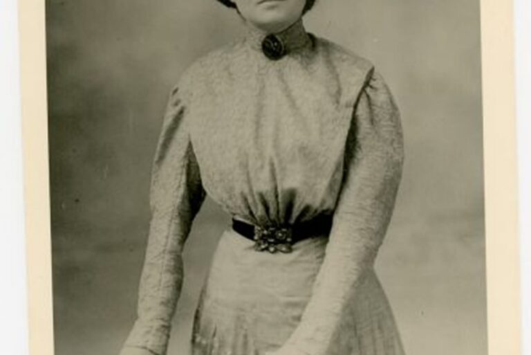 Vintage photo of a woman in a high-collared, long-sleeved dress, standing and looking at the camera.