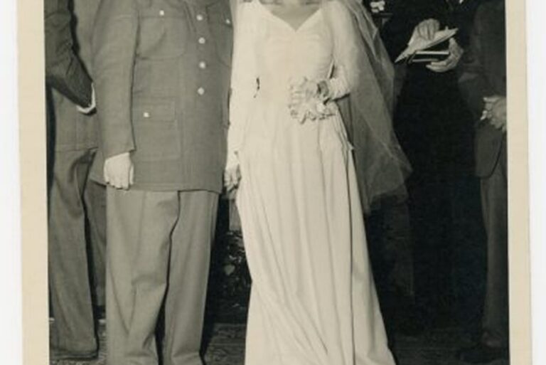 A bride in a long dress and veil stands next to a groom in a military uniform, both facing the camera.