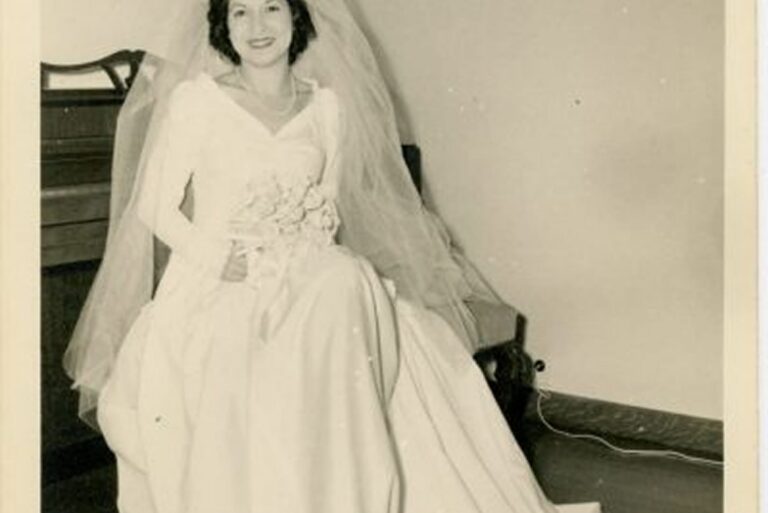 A bride in a vintage wedding dress and veil sits on a bench, smiling and holding a bouquet.