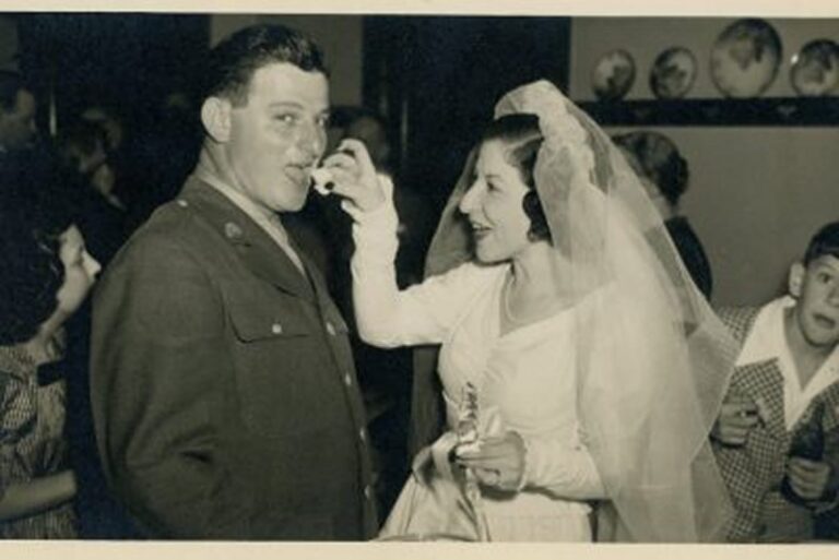 Bride in a wedding dress feeds cake to a groom in military uniform, with children smiling in the background.
