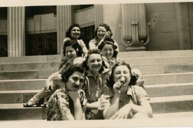 Seven women sit smiling and laughing together on stone steps in front of a building with columns.