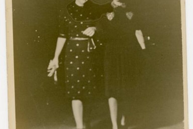 Two women in vintage dresses walk arm in arm, smiling at each other on a dimly lit street at night.