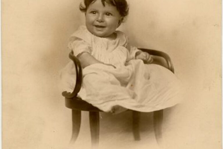 Smiling baby in a long white gown sits on a small curved wooden chair, vintage sepia photograph.