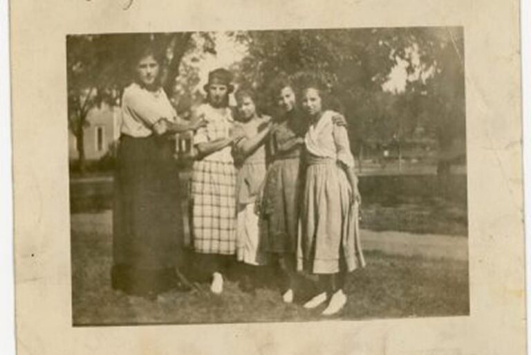 Five young women stand arm in arm outside, wearing dresses and white shoes, with trees and grass in the background.