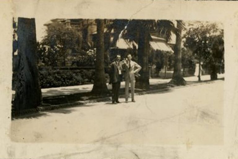 Two people in suits stand on a sunlit sidewalk lined with trees, with houses and foliage in the background.