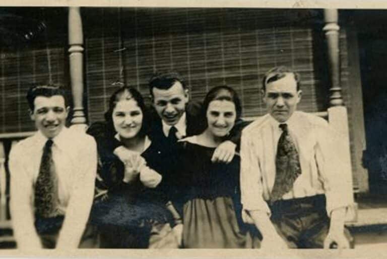 Five people pose closely together, smiling, in front of a porch with wooden railings and blinds in the background.