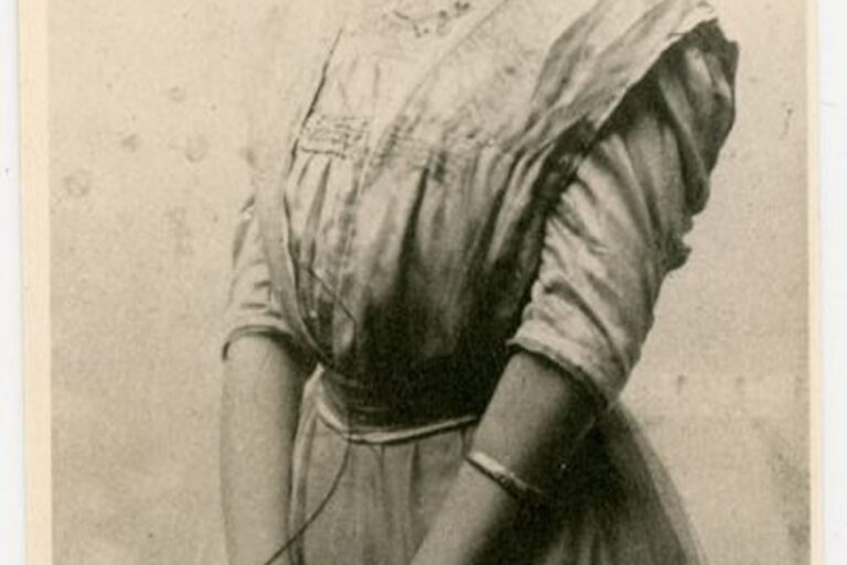 A young woman in a vintage dress poses for a sepia-toned portrait, hands clasped in front of her.